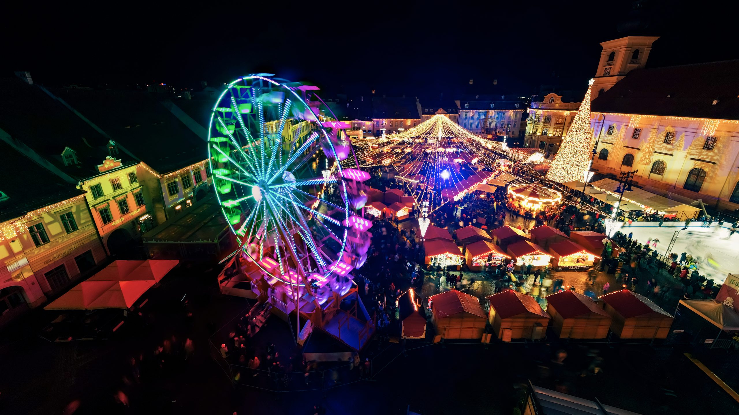 Aerial drone view of The Big Square in Sibiu at night, Romania. Old city centre decorated for Christmas. Ferris wheel, skating rink, old traditional buildings, people